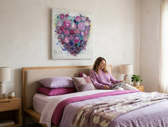 Quadro dipinto a mano con motivo a cuore di cerchi viola e rosa appeso in un interno moderno ed elegante sopra una consolle in legno con vaso e candela.